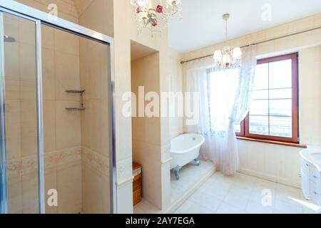 Bagno classico in toni beige. Vasca da bagno curva. Grande piastrella beige. Piastrelle bianche sul pavimento. Lampadario ritorto, elegante e costoso impianto idraulico Foto Stock