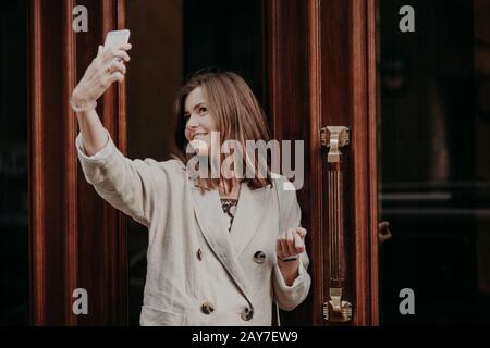 Donna adorabile fa selfie con smartphone, vestito con impermeabile, posa all'aperto, utilizza la tecnologia moderna, condivide le foto nei social network. Peo Foto Stock