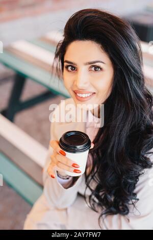 Colpo all'aperto di bella donna brunetta con un aspetto meraviglioso indossando abiti bianchi che tengono una tazza di carta di caffè delizioso. Femmina caucasica con Foto Stock