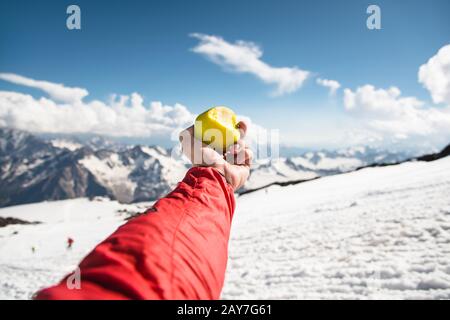 Una mano dell'uomo tiene una mela con un morso sullo sfondo di montagne innevate e neve sotto il piede. Foto Stock