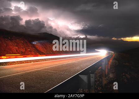 La strada di montagna con l'auto che passa per il tramonto sulla destra e un temporale con fulmine a sinistra è girato su una lunga expos Foto Stock