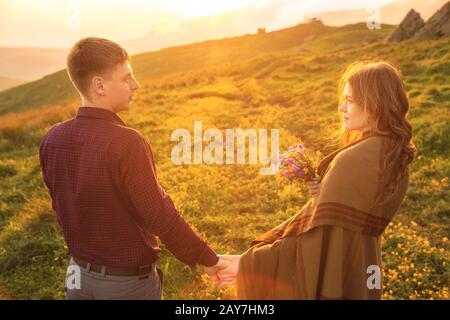 Una giovane coppia. Il ragazzo conduce una ragazza curly avvolto in un plaid con un bouquet di fiori al tramonto. Foto Stock