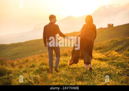 Una giovane coppia. Il ragazzo conduce una ragazza curly avvolto in un plaid con un bouquet di fiori al tramonto. Foto Stock