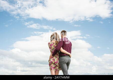 Vista dal retro di una giovane coppia si erge in un abbraccio e guarda fuori nella distanza contro il cielo con le nuvole. Foto Stock