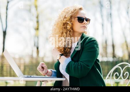 Giovane femmina graziosa con capelli biondi lanuginosi che indossano occhiali da sole seduti al sole davanti al laptop traducendo texte che guardano intorno notandola Foto Stock