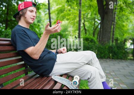 Lo skater da hippster a pelo lungo in un cappello si siede su una panchina e gira su una spinner Foto Stock