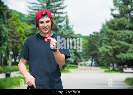 Un hipster dai capelli lunghi in un cappuccio si siede su una panchina e fa girare un fidget-spinner Foto Stock