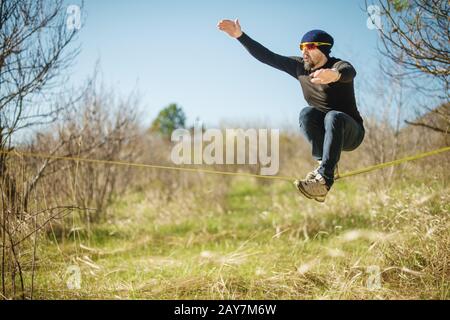 Un uomo all'età di sedersi in occhiali con un cappello e scarpe da ginnastica sul slackline, cattura l'equilibrio e gode la vita sulla natura i. Foto Stock