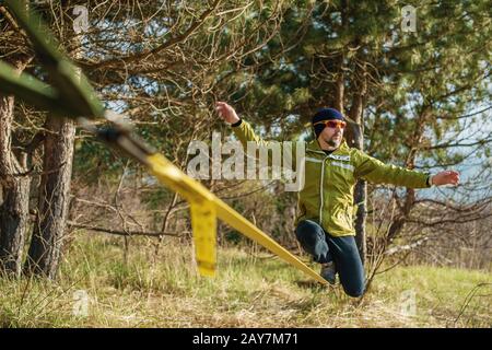 Un uomo all'età di sedersi in occhiali con un cappello e scarpe da ginnastica sul slackline, cattura l'equilibrio e gode la vita sulla natura i. Foto Stock