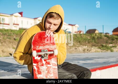 Primo piano di un adolescente vestito con una felpa con cappuccio jeans seduta in un parco skate e tenendo uno skateboard Foto Stock