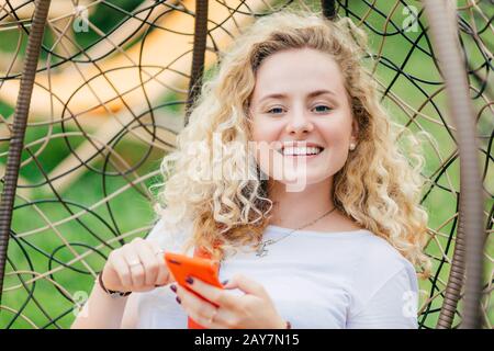 Giovane donna bionda soddisfatta con capelli ricci, ha sorriso positivo, usa il moderno cellulare per chattare con gli amici, can`t immaginare la sua vita senza mo Foto Stock