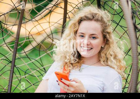 Femmina caucasica positiva con capelli ricci, si siede in sedia sospesa, ha sorriso amichevole, tipi di messaggi di testo su cellulare moderno, rende lo shopping online, Foto Stock