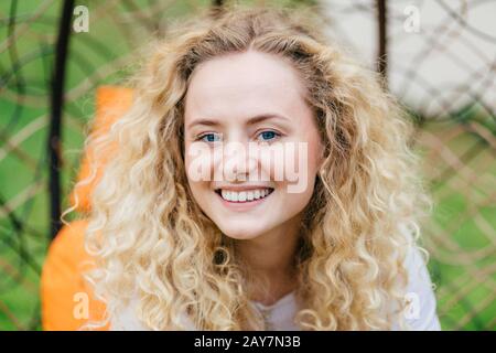 Foto di attraente femmina bionda curly con sorriso brillante, mostra denti bianchi pari, essendo in spirito alto, gode di buon riposo all'aperto, parla con l'amico. Foto Stock