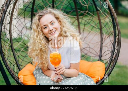 Foto di attraente bella femmina con capelli ricci chiaro con espressione allegra, recreats all'aperto in sedia sospesa, cocktail bevande gode di giornata soleggiata Foto Stock