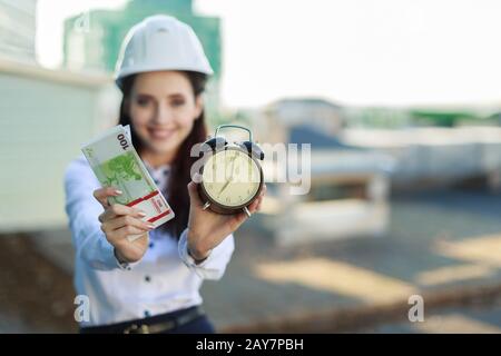Attraente donna d'affari in blusa bianca, casco e gonna nera stand sul tetto e tenere denaro e sveglia Foto Stock