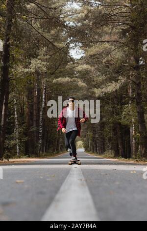 Un giovane ippopotere in una camicia con berretto e plaid sta cavalcando il suo longboard su una strada di campagna nella foresta Foto Stock