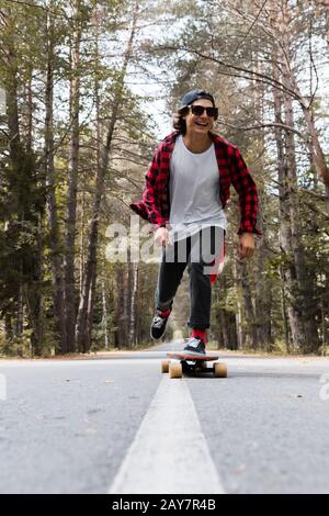 Un giovane hippster ridente in una maglia del berretto e del plaid sta cavalcando il suo longboard su una strada di paese nella foresta Foto Stock