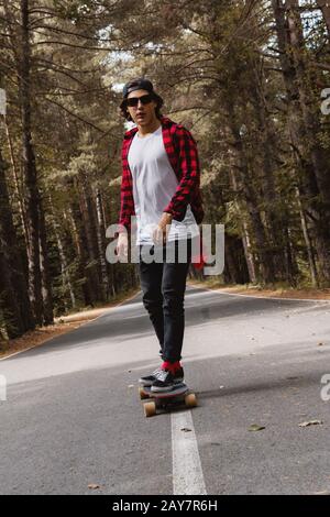 Un giovane ippopotere in una camicia con berretto e plaid sta cavalcando il suo longboard su una strada di campagna nella foresta Foto Stock