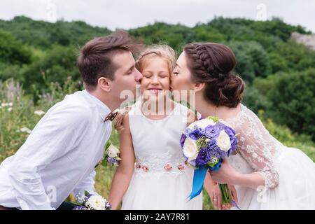 I giovani genitori in abiti da sposa baciano la loro giovane figlia in guance Foto Stock