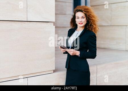 Bella giovane donna d'affari elegante in costume nero, avendo capelli ricci e attraente aspetto, tenendo tablet, leggendo le notizie su Internet whi Foto Stock