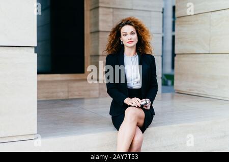 Attraente giovane donna che indossa abito nero formale, seduta gambe incrociate con dispositivo elettronico in attesa di business partner vicino al caffè. Splendida femmina Foto Stock