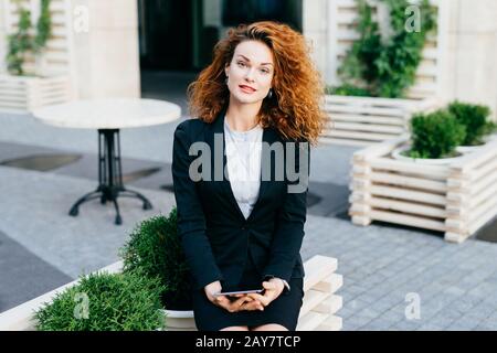 Giovane donna d'affari felice con capelli ricci, indossando blusa bianca e costume nero, seduto al ristorante all'aperto con tablet, utilizzando internet co gratuito Foto Stock