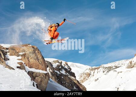 Uno sciatore professionista fa un salto-goccia da una scogliera alta contro un cielo blu che lascia un sentiero di polvere di neve nelle montagne Foto Stock