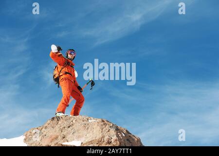 Ritratto di Uno Sciatore in una tuta arancione con uno zaino sulla schiena e gli sci sulle spalle in un casco si erge su un agai rock Foto Stock