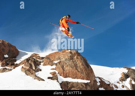 Uno sciatore professionista fa un salto-goccia da una scogliera alta contro un cielo blu che lascia un sentiero di polvere di neve nelle montagne Foto Stock