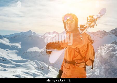 Ritratto di Uno Sciatore in una tuta arancione con uno zaino sulla schiena e gli sci sulle spalle in un casco si erge su un agai rock Foto Stock