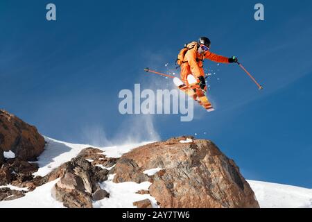 Uno sciatore professionista fa un salto-goccia da una scogliera alta contro un cielo blu che lascia un sentiero di polvere di neve nelle montagne Foto Stock