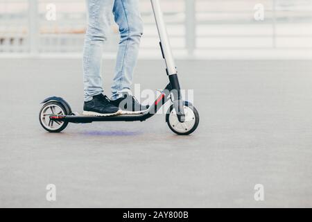 Gambe di un maschio sconosciuto in sneakers nere e jeans su scooter elettrico su asfalto urbano, gode di una giornata di sole. Persone e tempo libero concetto Foto Stock