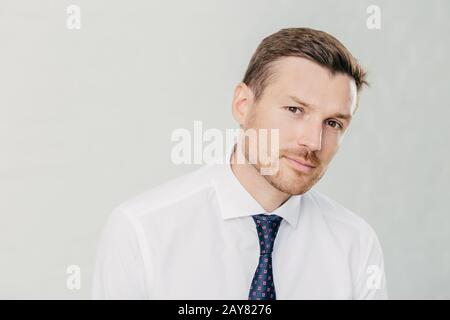 Il maschio sicuro guarda seriamente la fotocamera, ha occhi scuri e stoppia, vestito in elegante camicia bianca con cravatta, isolato su sfondo bianco. Uomo terribile Foto Stock