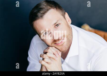 Ritratto di giovane attraente unshaven maschio caucasico con stoppie, ha un aspetto positivo, mantiene le mani sotto il mento, vestito con eleganti vestiti bianchi, isolat Foto Stock