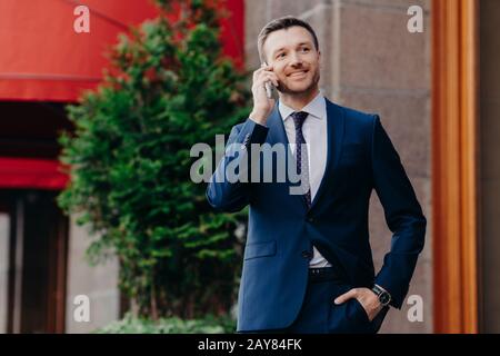 Ottimista uomo proprietario di buisness compagnia vestita in abito elegante, ha conversazione telefonica, consulta con operatore, guarda a distanza, ha felice Foto Stock