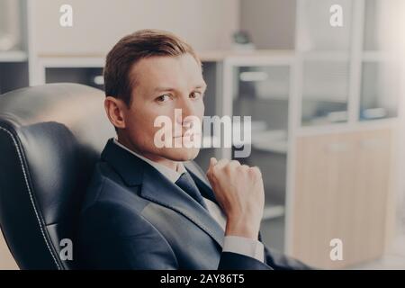 Il manager maschile professionale in abito formale, guarda direttamente alla macchina fotografica, riposa in sedia al posto di lavoro, confemples circa il nuovo progetto di business. Uomo elegante Foto Stock