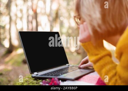 Un'immagine mock-up della mano di una donna che usa e digita su un laptop con un desktop nero vuoto su un prato plaid che giace nella foresta. Foto Stock