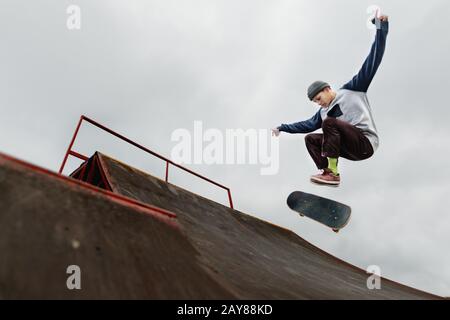 Teenager skateboarder in un cappello facendo un salto trucco su un tubo a metà su uno sfondo cielo nuvoloso Foto Stock