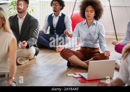 gruppo di colleghi di lavoro che meditano al lavoro, seduti sul pavimento con gambe incrociate e occhi chiusi. concetto moderno, informale, aziendale, di meditazione Foto Stock