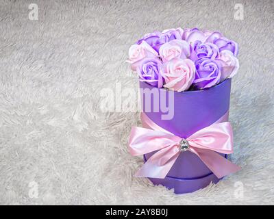 Rose rosa e viola in una scatola viola cilindrica legata con un nastro di seta rosa su uno sfondo di pelliccia bianca. Delicato regalo romantico per compleanno, donna Foto Stock