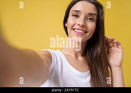 Piacevole ragazza attraente che fa selfie in studio e ridendo. Giovane donna dall'aspetto buono con capelli marroni che scattano l'immagine dell'erigella Foto Stock