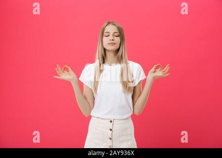 Bionda beauiful giovane femmina rende mudra segno, si rilassa dopo dura giornata di lavoro, mantiene gli occhi chiusi, pratica yoga contro la schiena rosa Foto Stock