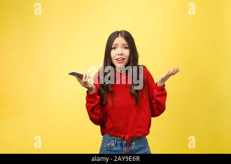 Ritratto arrabbiato giovane donna asiatica delude al telefono cellulare mentre in piedi su sfondo giallo Foto Stock