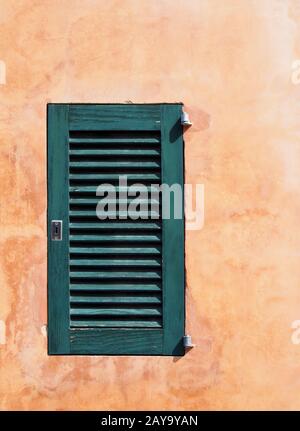 una piccola serranda di legno dipinta di verde su una vecchia parete ruvida di colore arancio ocra in sole brillante Foto Stock