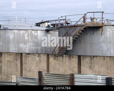 vecchi serbatoi di stoccaggio industriale in acciaio con scale arrugginite e passerelle dietro un recinto di ferro ondulato shabby e muro Foto Stock