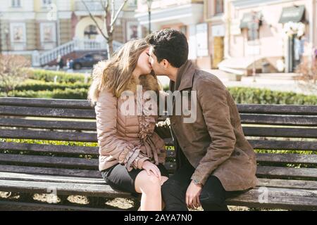 Giovane coppia multirazziale, maschi e femmine eterosessuali studenti innamorati. Una data in un parco cittadino su una panchina di legno. Giovane brunetta Foto Stock