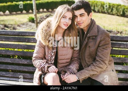 Giovane coppia multirazziale, maschi e femmine eterosessuali studenti innamorati. Una data in un parco cittadino su una panchina di legno. Giovane brunetta Foto Stock