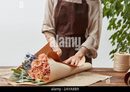 Donna fiorista sta avvolgendo il bouquet di rosa fresco in una carta decorativa su un tavolo di legno su uno sfondo muro chiaro. Procedere passo dopo Foto Stock