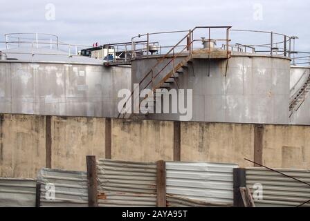 vecchi serbatoi industriali in metallo con scale di ispezione arrugginite e valvole circondate da una recinzione e da una parete in acciaio ondulato Foto Stock