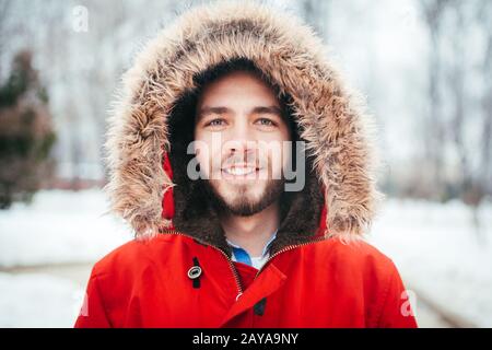 Ritratto, primo piano di un giovane uomo vestito elegantemente sorridente con una barba vestita con una giacca rossa d'inverno con cappuccio e pelliccia su h Foto Stock